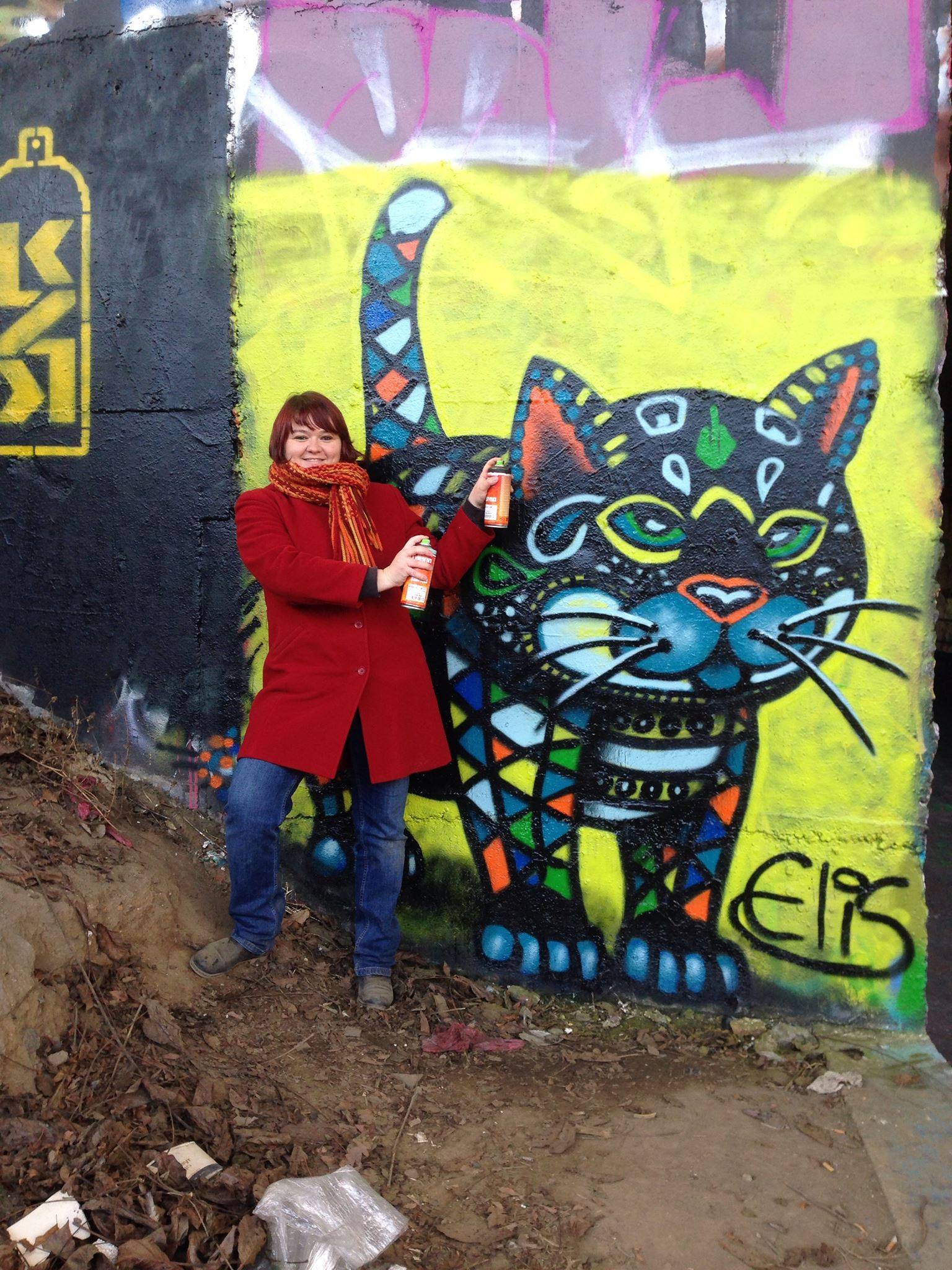 Eliška Hesslerová se dvěma sprejemi před velkým graffiti barevné kočky podepsaným Elis